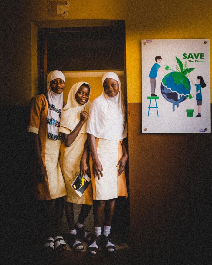 Three schoolgirls in uniform pose beside an environmental poster indoors.